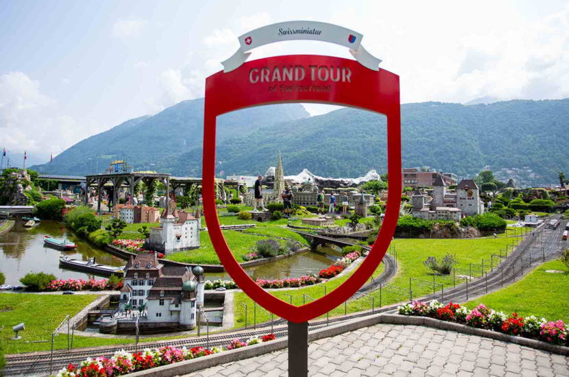 Zoom: Der rote Grand Tour of Switzerland Fotopoint eröffnet den Blick auf die Modelllandschaft der Swissminiatur in Melide mit Miniaturgebäuden, Bahnstrecken und der Bergkulisse des Luganersees.