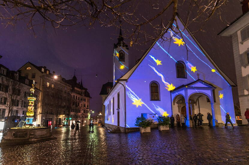 Zoom: Die historische Kirche in Luzern wird beim Lichtfestival in ein funkelndes Sternenmeer verwandelt. Blaue Lichtstrahlen und goldene Sterne schaffen eine traumhafte Kulisse in der Altstadt und machen das Gebäude zu einem beliebten Fotospot.