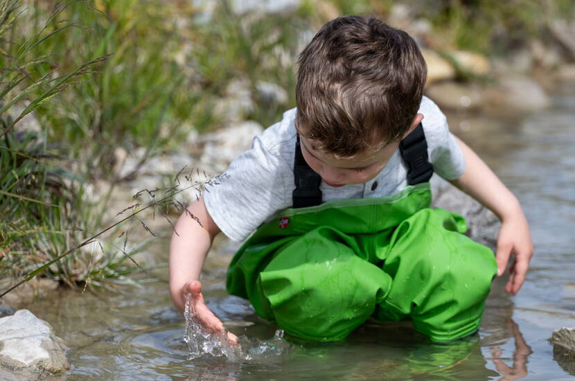Zoom: Kleinkind kniet im Bach und spielt im Wasser mit wasserdichter grüner Rægni Wathose - perfekt für Naturerlebnisse