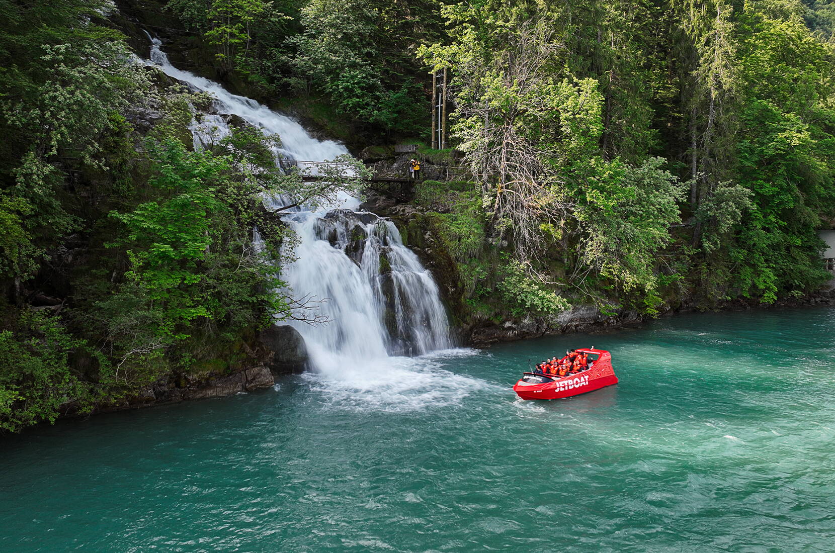 Un Jetboat rouge se dirige vers les chutes du Giessbach, qui dévalent plusieurs niveaux rocheux jusqu’au lac. Les passagers profitent d’une vue impressionnante sur la cascade, la forêt et les eaux turquoise du lac de Brienz.