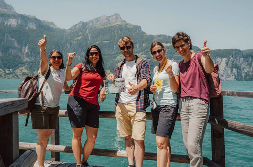 Zoom: Six personnes jubilantes sur ponton en bois du lac des Quatre-Cantons turquoise avec toile de fond montagneux dramatique célèbrent la résolution réussie de l'affaire criminelle Tatort Tell