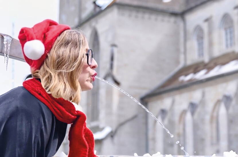 Zoom: Une personne pose devant une fontaine historique, donnant l’impression que le jet d’eau fait partie de la scène – le tout agrémenté d’un bonnet et d’une écharpe rouges pour une touche festive.
