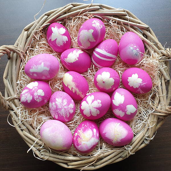 Un panier rond rempli d’œufs de Pâques teints en rose et décorés de motifs floraux et végétaux clairs.