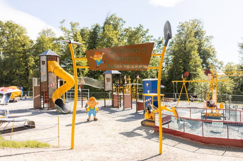 Schongis Kinderbaustelle im Schongiland Aussenaufnahme eines Spielbereichs mit dem Eingangsschild «Schongis Kinder-Baustelle». Verschiedene Spielgeräte und bunte Elemente sind sichtbar, umgeben von Bäumen.