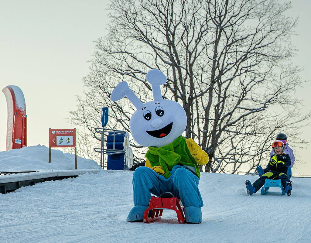 Fröhliches Maskottchen und Kinder beim Schlitteln auf der präparierten Piste der Bodmi Arena in Grindelwald. Familienfreundliches Wintervergnügen für Gross und Klein im Berner Oberland.
