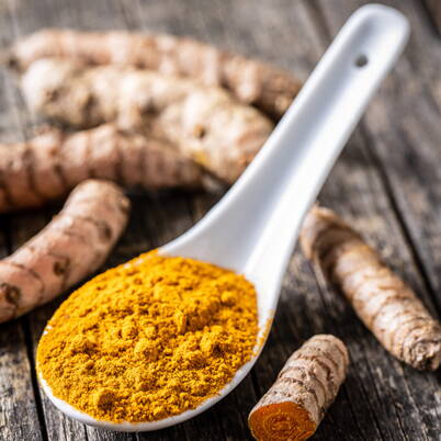 Une cuillère blanche remplie de poudre de curcuma jaune repose sur une table en bois, accompagnée de racines de curcuma fraîches.