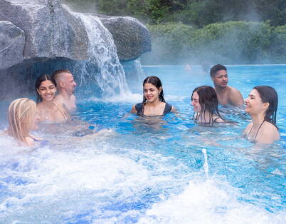 Des visiteurs profitent de l’eau bouillonnante et de la cascade dans le bassin extérieur chauffé, entouré de nature.
