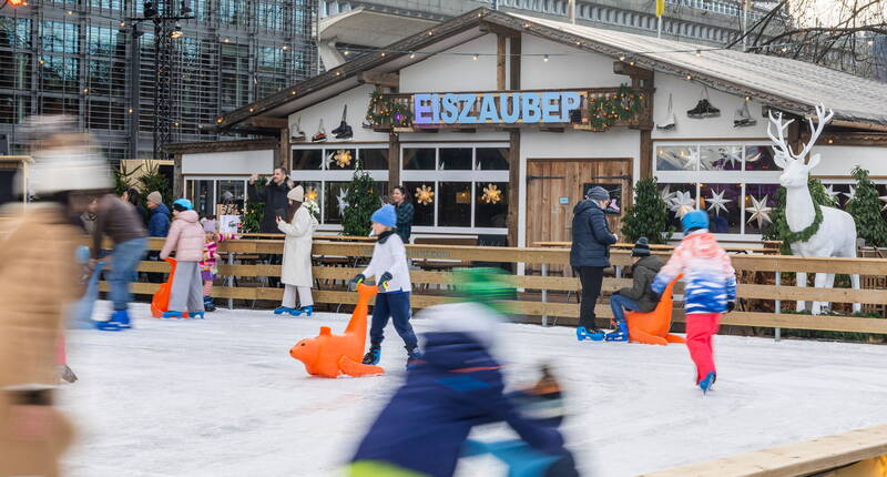 Kinder und Familien beim kostenlosen Schlittschuhlaufen auf der Eisfläche des Eiszauber Luzern mit Chalet im Hintergrund