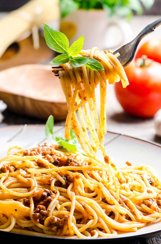 Spaghetti Bolognese mit frischen Kräutern Ein Teller Spaghetti Bolognese, garniert mit frischem Basilikum. Die Pasta wird mit einer Gabel elegant angehoben, im Hintergrund sind Tomaten und Küchenutensilien zu sehen.