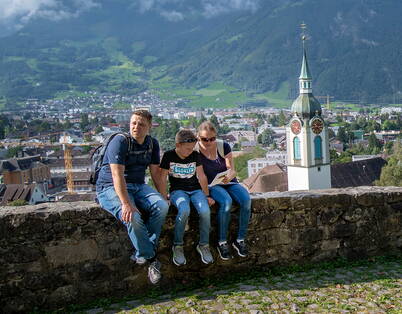 Eine Familie sitzt auf einer Steinmauer. Sie versuchen eines der Rätsel der Schnitzeljagt zu lösen. Im Hintergrund sieht man die Kirche und Altdorf.