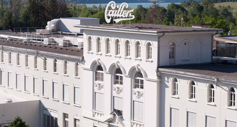 La chocolaterie historique Maison Cailler à Broc, avec le logo Cailler sur le toit et des arbres en arrière-plan.