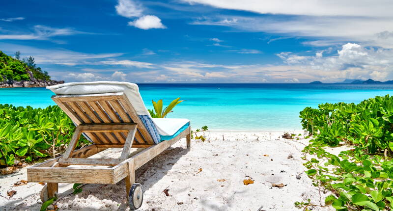 Sonniger Strandabschnitt mit Holzliege vor intensiv blauem Wasser.
