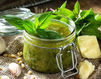 Ein Glas mit frisch zubereitetem Kräuterpesto, umgeben von Basilikum, Knoblauch, Pinienkernen und Parmesan.