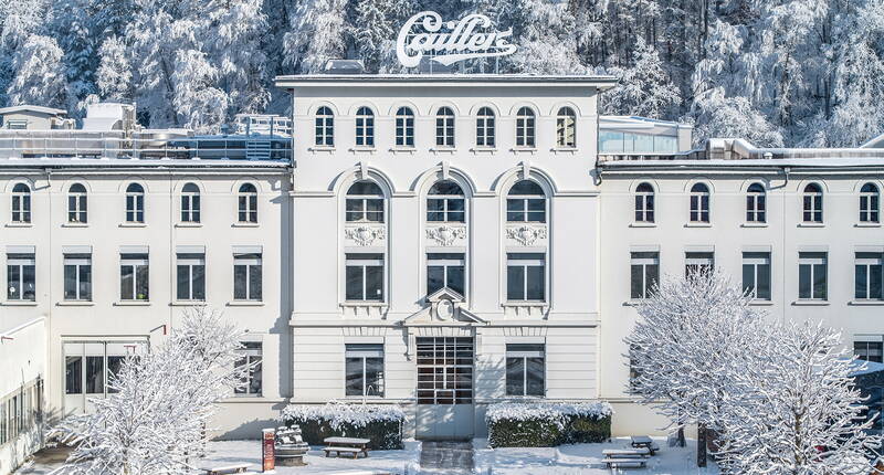 Die historische Schokoladenfabrik Maison Cailler in Broc, umgeben von verschneiter Landschaft und einem kleinen Zug im Vordergrund.