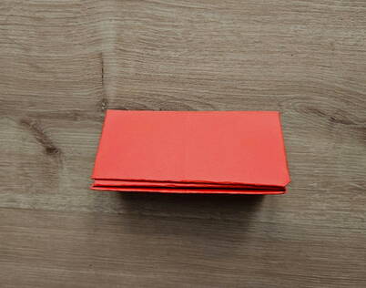 DIY Papierportemonnaie – Zusammengefaltete Mittelpartie Das rote Papier wurde längs zu einem kompakten Rechteck zusammengefaltet. Dieser Schritt verdichtet die Struktur des DIY Papierportemonnaies und bildet die Basis für die spätere Fächerform. Die sauberen, parallel verlaufenden Kanten sorgen dafür, dass das Origami‑Portemonnaie stabil bleibt und sich später korrekt öffnen und schliessen lässt.