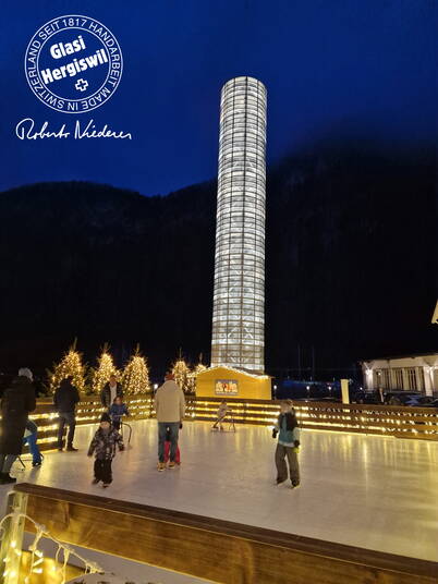 Eislauffeld der Glasi Hergiswil Zu sehen ist ein Eislaufplatz bei Nacht mit beleuchteten Tannenbäumen und einem hohen, hell erleuchteten Turm im Hintergrund.