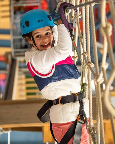 Una persona è assicurata con imbragatura e corda di sicurezza su un percorso di corde. Indossa un casco blu e si tiene a una corda, mentre i moschettoni garantiscono la sicurezza. Sullo sfondo si vedono elementi in legno e altre parti del percorso.