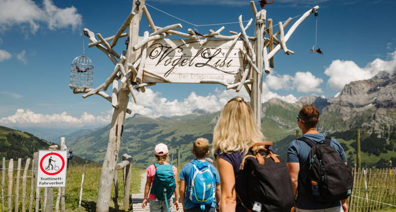 Familie am Eingang des Vogellisi-Erlebniswegs auf dem VogellisiBerg in Adelboden. Der beliebte Familienausflug im Berner Oberland verbindet Natur, Wandererlebnis und die Geschichte des Vogellis.