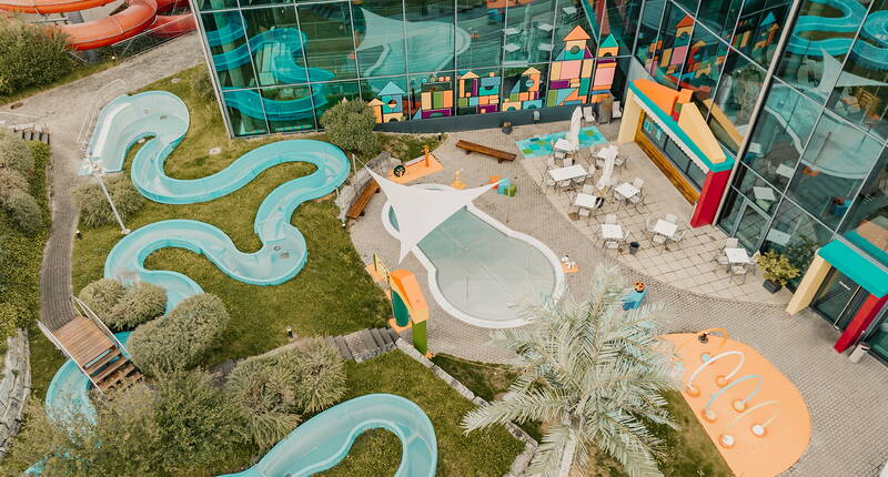 Vista dall’alto sull’area esterna colorata con canale d’acqua curvo, zone gioco e spazi relax.