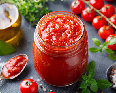 Ein geöffnetes Glas voller aromatischer Tomatensauce, umgeben von frischen Tomaten, Kräutern und Gewürzen – perfekt zur weiteren Verarbeitung oder zum Haltbarmachen.