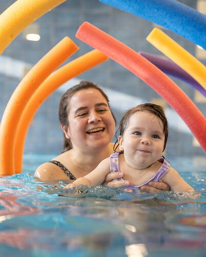 Ein kleines Kind wird im Wasser gehalten, während bunte Schwimmnudeln wie ein farbenfroher Tunnel über dem Wasserbogen angeordnet sind. Die Umgebung ist ein Hallenbad mit ruhiger Wasseroberfläche und blauen Fliesen.
