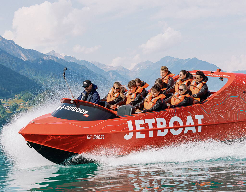 Un Jetboat rosso percorre il Lago di Brienz ad alta velocità, sollevando spruzzi potenti ai lati. I passeggeri siedono nella parte anteriore mentre l’imbarcazione si dirige verso il largo. Il paesaggio montano dell’Oberland bernese crea uno sfondo spettacolare.