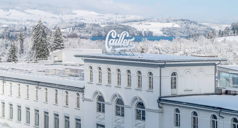 Die Schokoladenfabrik Maison Cailler in Broc mit winterlicher Aussicht auf die umliegenden Hügel und Wälder.
