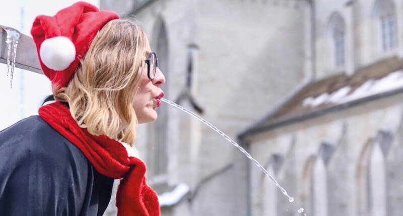 Una persona posa davanti a una fontana storica, facendo sembrare il getto d’acqua parte della scena – il tutto con cappello e sciarpa rossi per un tocco natalizio.