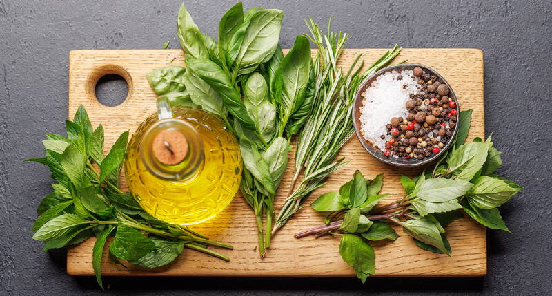 Verschiedene frische Kräuter, grobes Salz und Pfefferkörner liegen auf einem Holzbrett, ergänzt durch eine Karaffe mit aromatischem Kräuteröl.