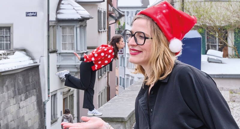 Due persone realizzano una foto creativa: una è sul muretto con il cappello di Natale, l’altra sembra appesa alla facciata – un effetto divertente che sembra un bacio da lontano.
