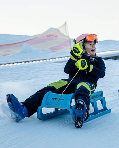 Una persona è seduta su una slitta di plastica blu e scivola su una pista innevata preparata. Indossa un casco, occhiali da sci e guanti ben visibili, mentre si tiene a una corda per stabilità. Sullo sfondo si vedono cumuli di neve e una rete di sicurezza.