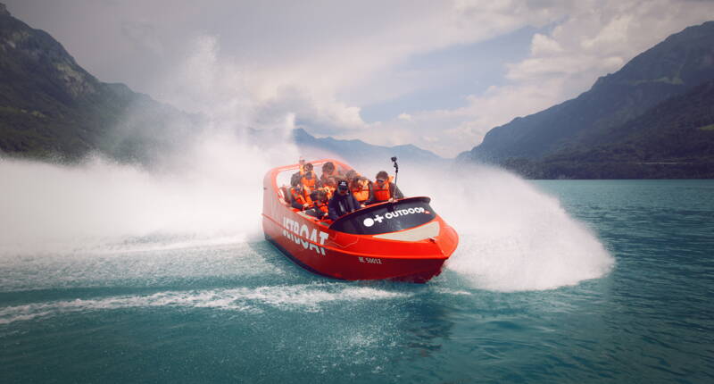 Un Jetboat orange glisse à grande vitesse sur les eaux turquoise du lac de Brienz. D’imposantes gerbes d’eau s’élèvent des deux côtés tandis que les passagers profitent de l’expérience. En arrière‑plan se dresse le paysage montagneux de l’Oberland bernois sous un ciel légèrement nuageux.