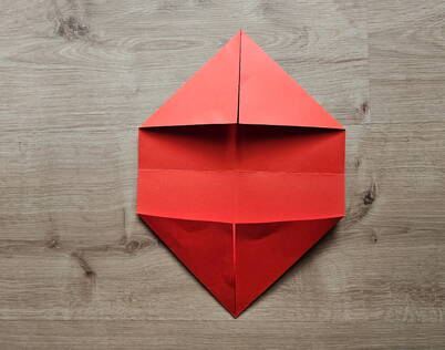 Papierportemonnaie falten – Obere und untere Spitzen zu Dreiecken formen Die obere und untere Spitze des roten Papiers werden zur Mittellinie gefaltet und bilden zwei gleichmässige Dreiecke. Dieser Schritt legt die Grundform für das spätere DIY Papierportemonnaie fest und schafft die ersten stabilen Kanten, die für das Origami‑Modell wichtig sind.