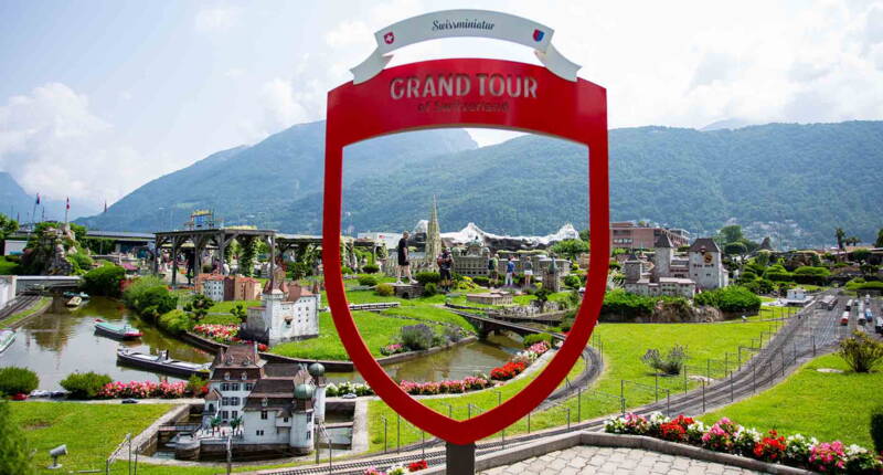 Der rote Grand Tour of Switzerland Fotopoint eröffnet den Blick auf die Modelllandschaft der Swissminiatur in Melide mit Miniaturgebäuden, Bahnstrecken und der Bergkulisse des Luganersees.