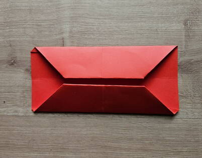 Papierportemonnaie falten – Längsseiten zur Mitte geklappt Die langen Seiten des roten Papiers wurden zur Mittellinie hin gefaltet und ergeben eine symmetrische, schmale Form. Dieser Schritt bildet die innere Struktur des DIY Papierportemonnaies und definiert die spätere Öffnung des origamiartigen Geldbeutels. Saubere Faltkanten sorgen dafür, dass das Portemonnaie später stabil und gleichmässig bleibt.