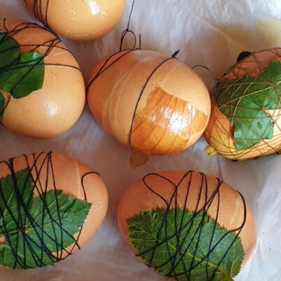 Diverse uova marrone chiaro giacciono sulla carta e sono ricoperte di foglie verdi. Un filo nero è avvolto stretto e in croce attorno a ogni uovo per mantenere le piante in posizione durante la colorazione naturale. La superficie delle uova è leggermente lucida.
