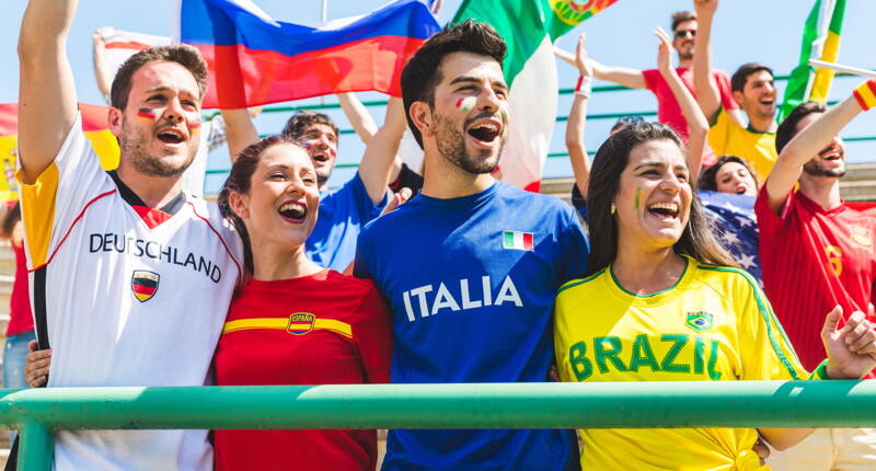 Persone con diverse maglie di squadre nazionali in una zona dello stadio che sollevano bandiere e celebrano un evento calcistico. Focus sull’abbigliamento dei tifosi e sull’atmosfera calcistica.