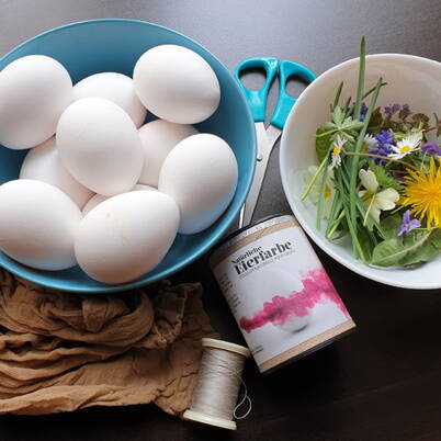 Weisse Eier, Kräuter, Blätter und eine Nylonstrumpfhose liegen bereit – die Materialien für natürlich gefärbte Ostereier mit Pflanzenmustern.