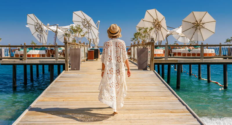 Passerelle en bois au‑dessus d’une eau claire avec vue sur un resort côtier sous un ciel lumineux.