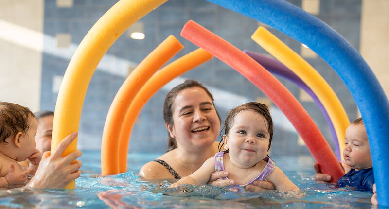 Bambini si divertono con tubi galleggianti colorati, guidati da adulti. Un’attività che aiuta a familiarizzare con l’acqua in modo giocoso.