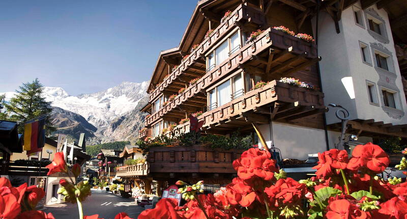Un hôtel en bois avec des balcons fleuris dans un village alpin pittoresque. En arrière-plan, des sommets enneigés dominent le paysage, tandis que des géraniums rouges apportent une touche de couleur au premier plan.