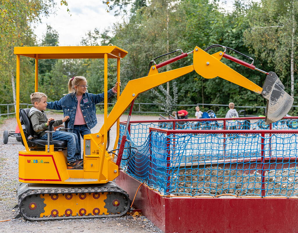 Kinderbagger auf der Baustelle im Schongiland Zwei Kinder bedienen gemeinsam einen gelben Mini‑Spielbagger in einem abgegrenzten Spielbereich. Im Hintergrund sind weitere Spielangebote und Bäume sichtbar.
