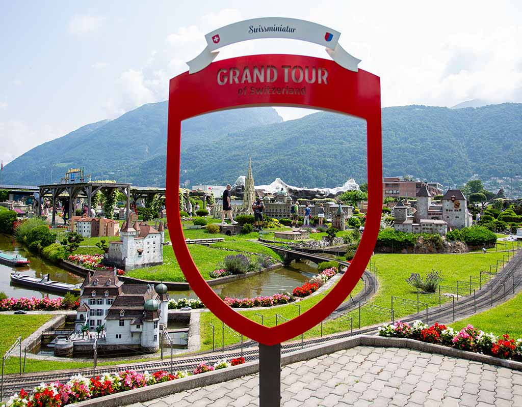 Grand Tour of Switzerland Fotopoint in der Swissminiatur Der rote Grand Tour of Switzerland Fotopoint eröffnet den Blick auf die Modelllandschaft der Swissminiatur in Melide mit Miniaturgebäuden, Bahnstrecken und der Bergkulisse des Luganersees.