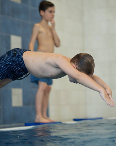 Un bambino si tuffa dal bordo della piscina con le braccia tese e il corpo dritto. Sullo sfondo, un altro bambino è in piedi su un tappetino. La scena si svolge in una piscina coperta con piastrelle blu e bianche sulla parete.
