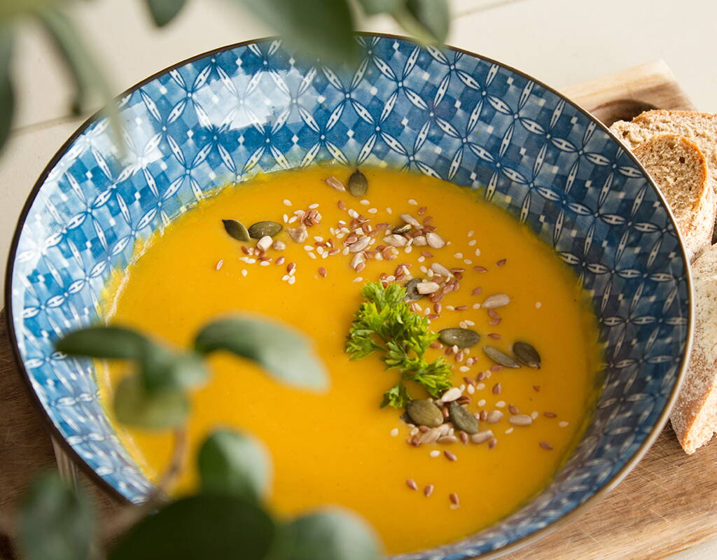 Cremige Kürbissuppe in einer dekorativen blauen Schale, garniert mit Kernen und frischen Kräutern. Die warme Farbgebung und das knusprige Brot machen Lust auf gemütliche Herbstgerichte.