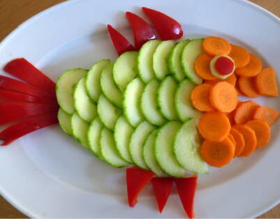 Fisch aus frischem Gemüse Auf dem weissen Teller ist ein Fisch aus Rüebli und Gurkenscheiben sowie roten Peperoni als Flossen und Schwanzflosse gelegt.