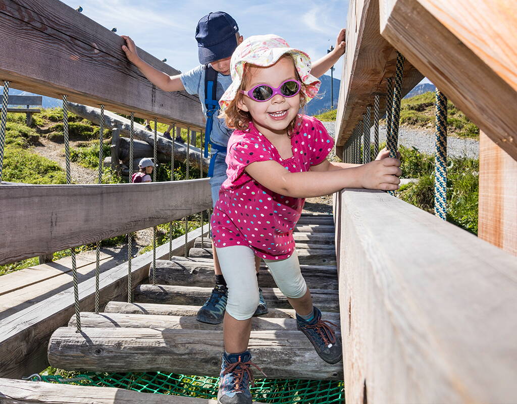 Kleines Mädchen balanciert über einen Parcours aus hängenden Baumstämmen auf dem Murmeli-Trail, im Hintergrund ein älterer Junge