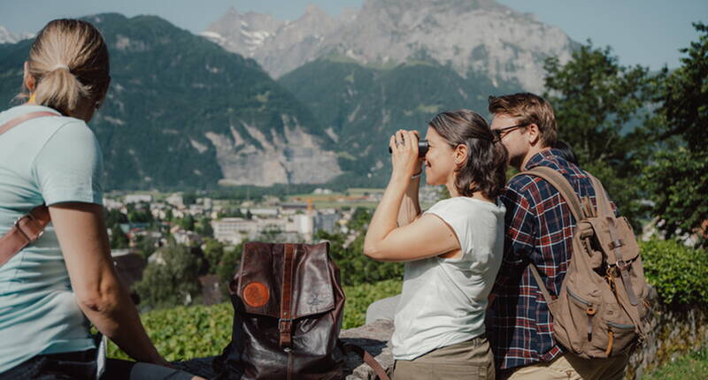 Un couple se tient devant un mur. Le sac à dos de jeu est devant eux et la femme cherche des indices avec des jumelles.