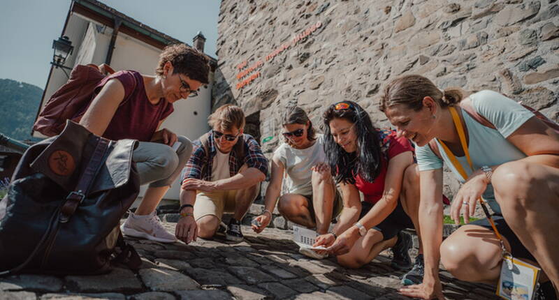 Squadra di investigatori inginocchiata su selciato storico davanti a muro di pietra ad Altdorf esamina insieme carte di gioco e indizi del giallo Tatort Tell