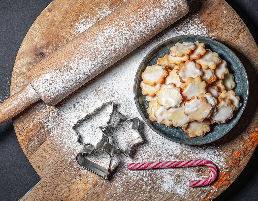 Auf einem runden Holzbrett liegen ein Nudelholz, Ausstechformen, eine Zuckerstange und ein Teller mit Mailänderli, die mit Zuckerguss verziert sind. Das Brett ist mit Puderzucker bestäubt – bereit für die Weihnachtsbäckerei.